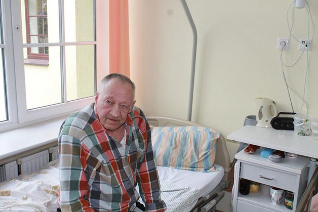elderly man sitting at corner of his bed with a tired look on his face